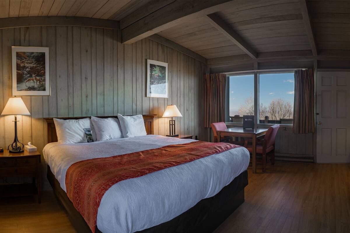 A bedroom with a large bed, two bedside tables, a desk, and a chair. There are two windows with curtains, and the room has wooden walls and ceiling.