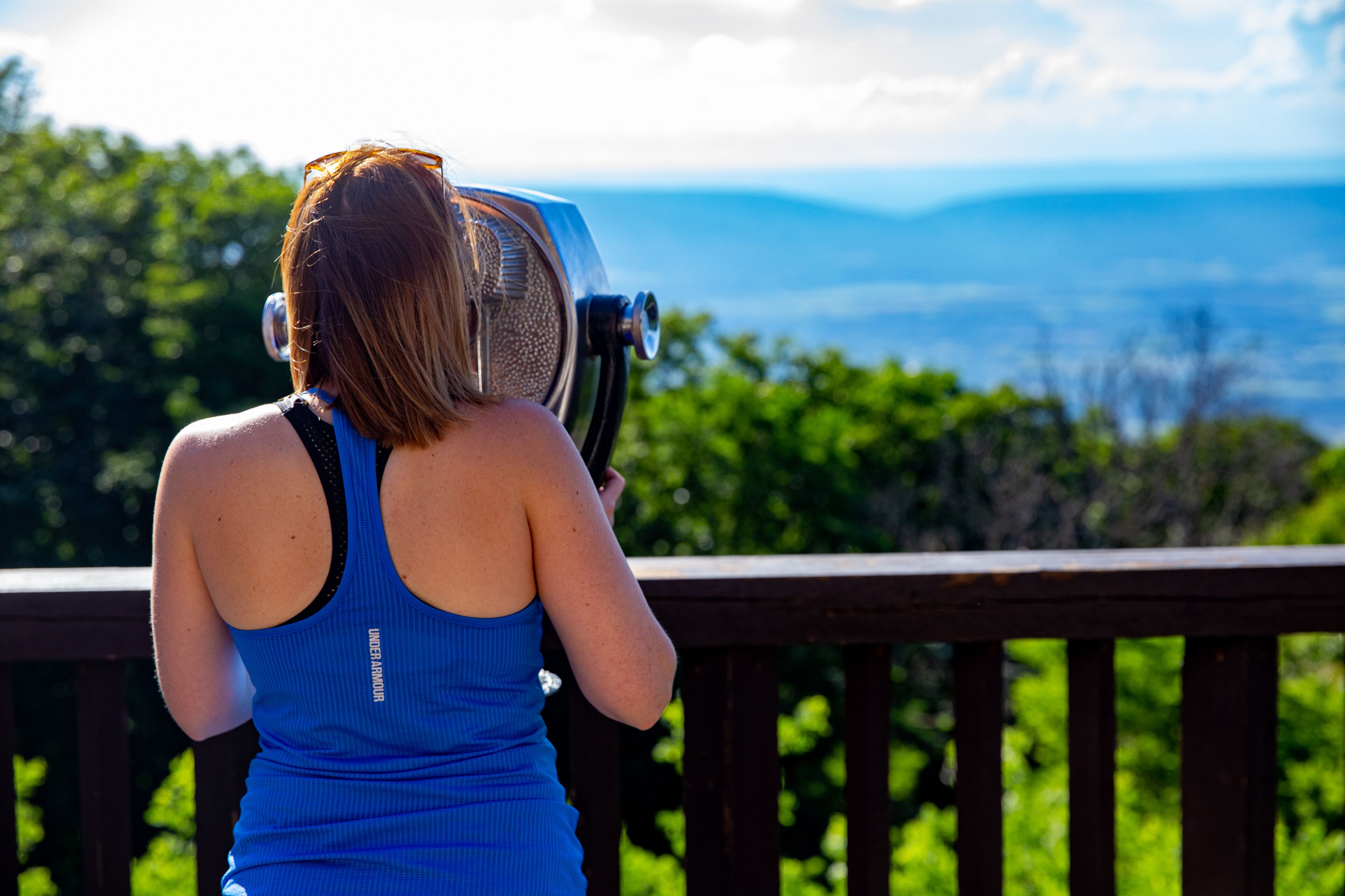 Eine Person beobachtet mit Fernglas eine gebirgige Landschaft von einer Holzdielen-Terrasse aus.