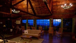 Une pièce spacieuse avec des poutres en bois, un canapé, des fauteuils et une vue dégagée grâce à de grandes fenêtres.