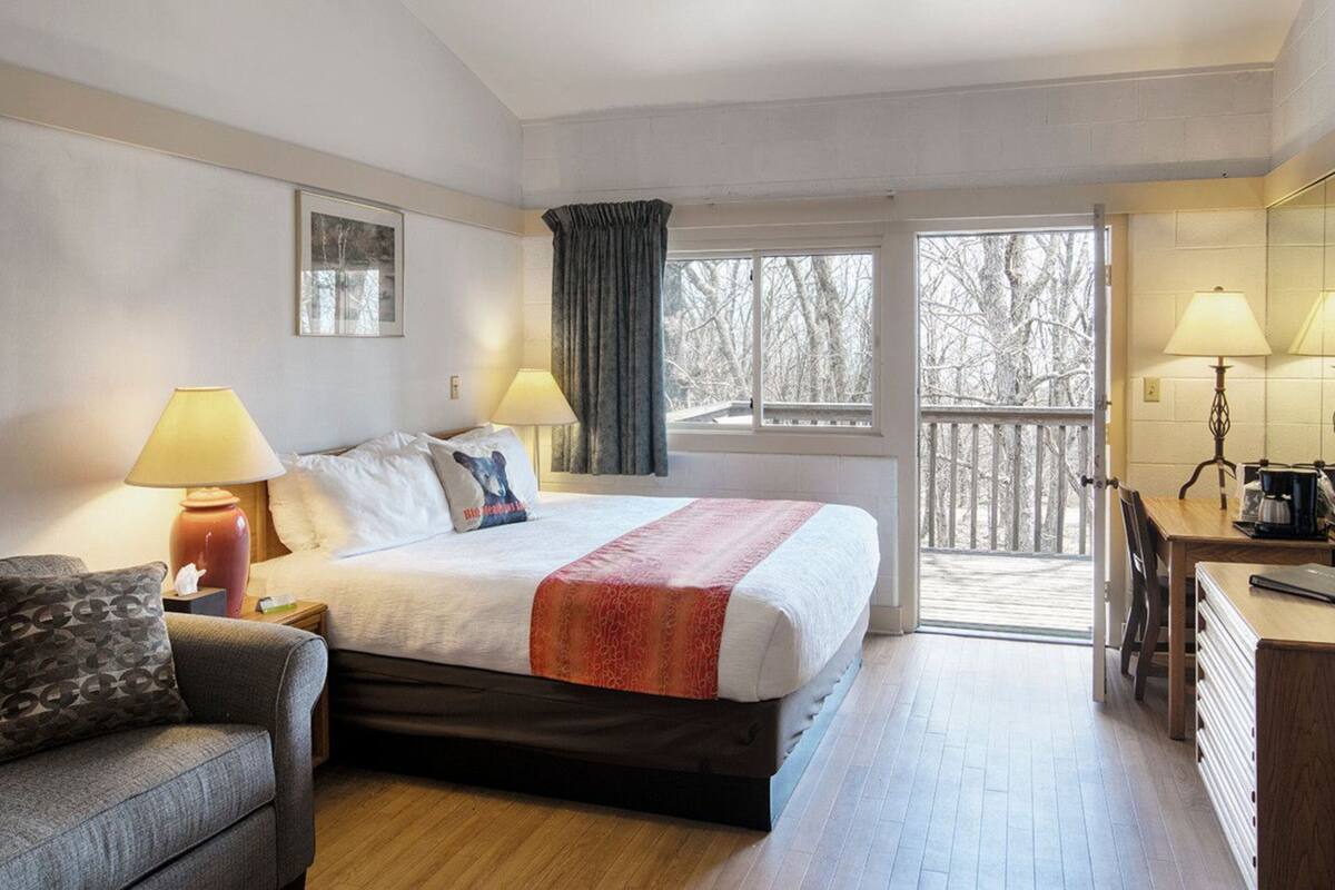 A hotel room with a bed, two bedside lamps, a sofa, a desk with a chair, and a view of trees through the window.