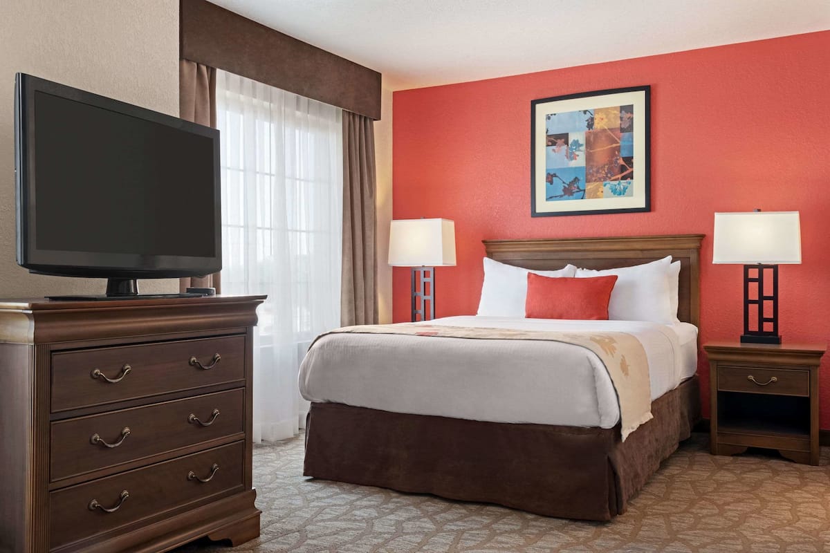 A hotel room with a large flat-screen TV on a dresser, a bed with white and red bedding, two bedside lamps, and a framed artwork on the wall.