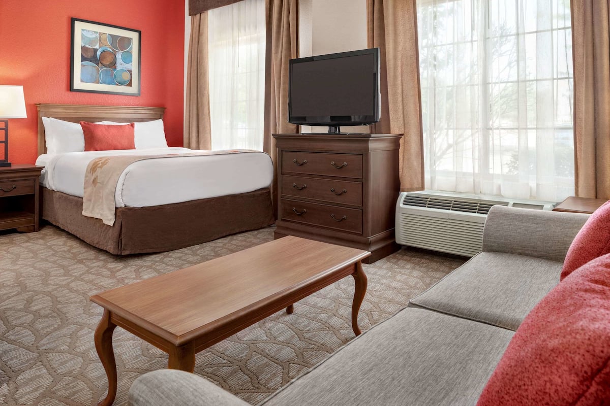 A hotel room with a bed, a TV on a stand, a wooden table, a sofa, and a window with curtains.