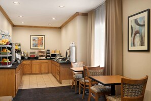 A hotel kitchen with wooden cabinets, a dining area with two chairs, a table, and a counter with various food items and appliances.