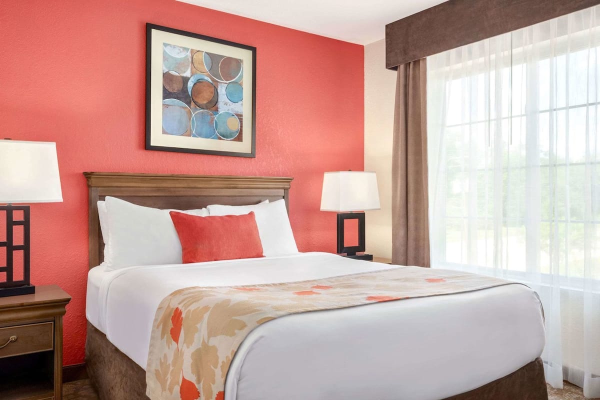 A hotel room with a bed, two bedside tables with lamps, a framed abstract painting, and a large window with sheer curtains.