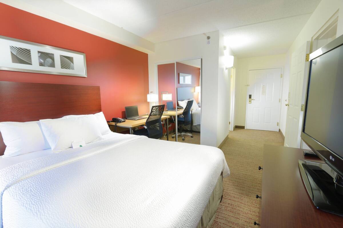 Et hotelværelse med en stor seng, et skrivebord med en computer, et fjernsyn og et spejl.