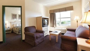 A hotel room with a bed, two armchairs, a small table, a TV, and a window with curtains. - Allure Suites (Fort Myers)
