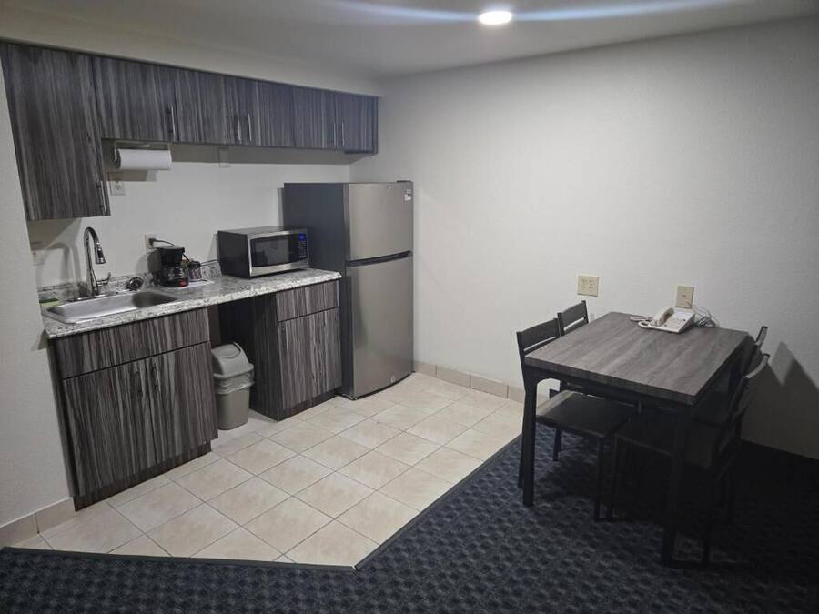 A compact kitchen with dark wood cabinets, a stainless steel refrigerator, a microwave, and a small sink. There is a dining table with chairs and a small trash bin.