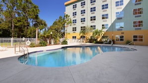 A swimming pool with a curved edge, surrounded by a concrete deck. There are steps leading into the pool, and a building with multiple windows and balconies in the background. - Allure Suites (Fort Myers)
