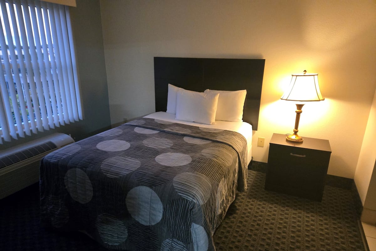 Habitación de hotel con cama, mesita de noche, lámpara y ventana con persianas.