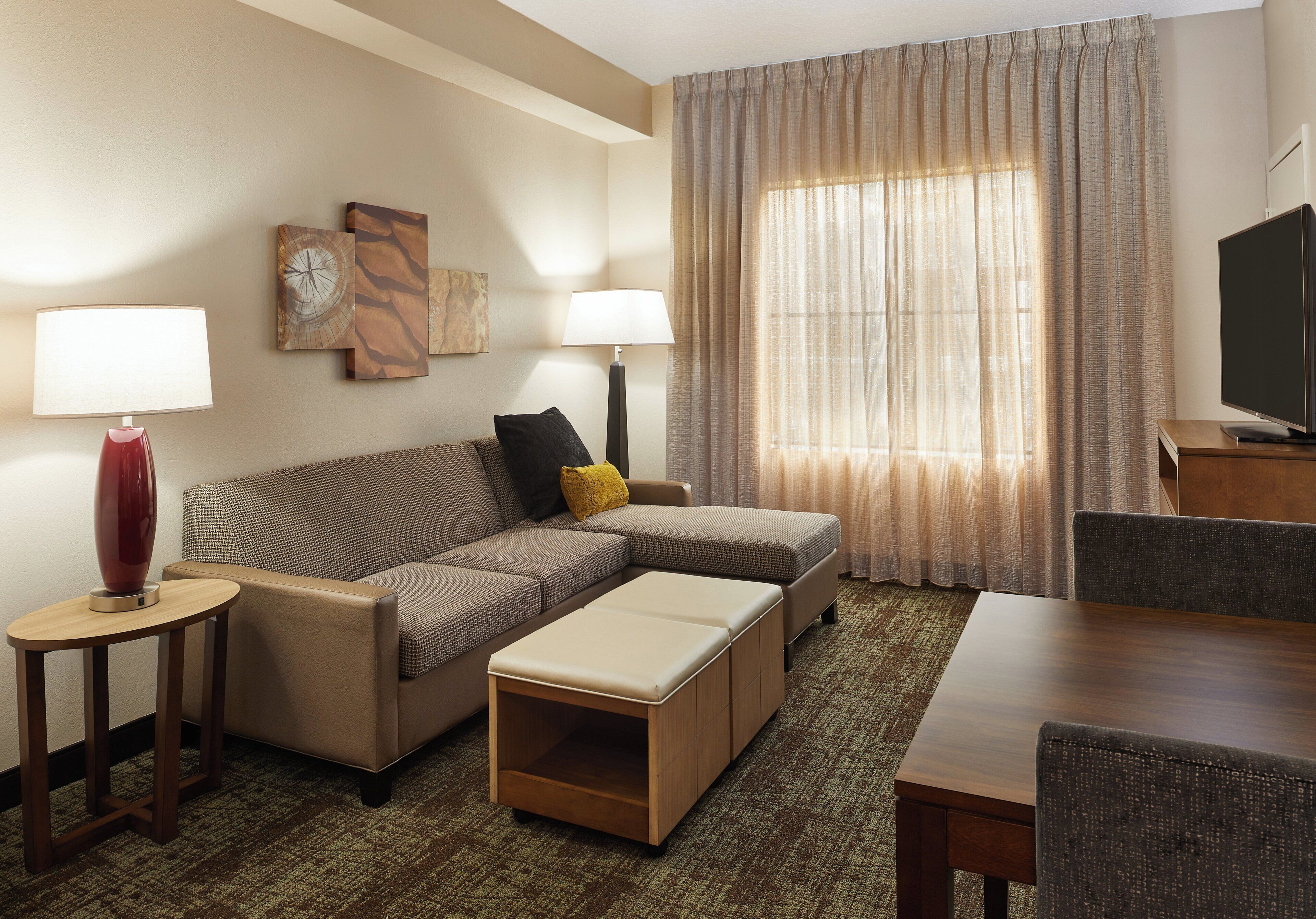 A hotel room with a sectional sofa, a small table, a TV, and a large window with curtains.