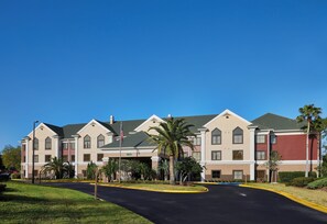 A multi-story hotel with a central courtyard, featuring a flagpole, palm trees, and a clear blue sky. - Staybridge Suites Orlando Airport South by IHG (Orlando)