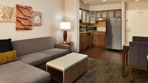 A modern living room with a sectional sofa, a small table, and a lamp. A kitchen is visible in the background with wooden cabinets and stainless steel appliances. - Staybridge Suites Orlando Airport South by IHG (Orlando)