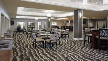 Uma sala de jantar de hotel com várias mesas e cadeiras, um tapete estampado e um teto com iluminação embutida.
