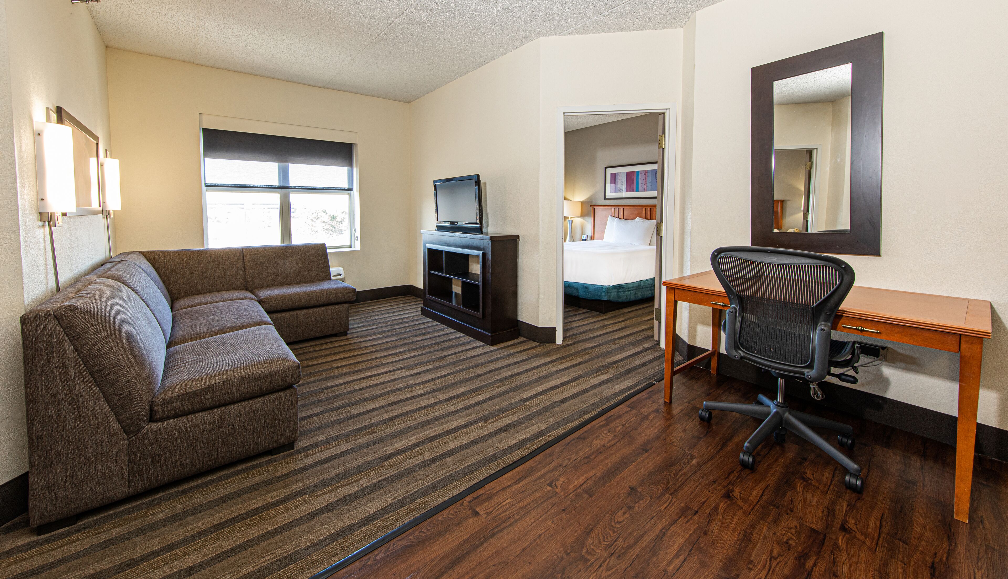 A hotel room with a sectional sofa, a TV stand, a desk with a chair, and a bed.