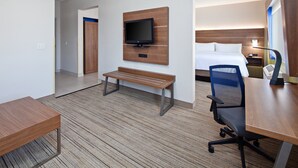 Une chambre d’hôtel avec un bureau en bois, un banc, un téléviseur fixé au mur, un lit et une table de chevet avec une lampe.