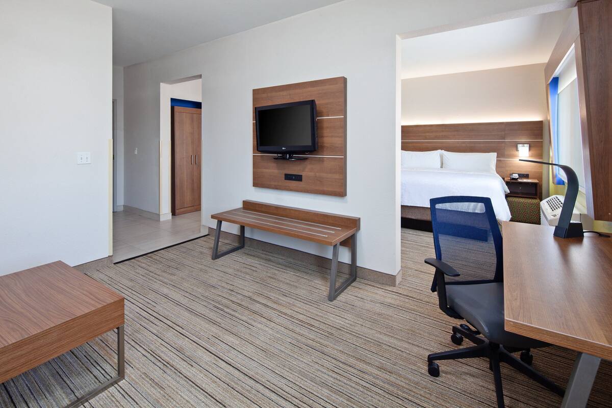 Une chambre d’hôtel avec un bureau en bois, un banc, un téléviseur fixé au mur, un lit et une table de chevet avec une lampe.