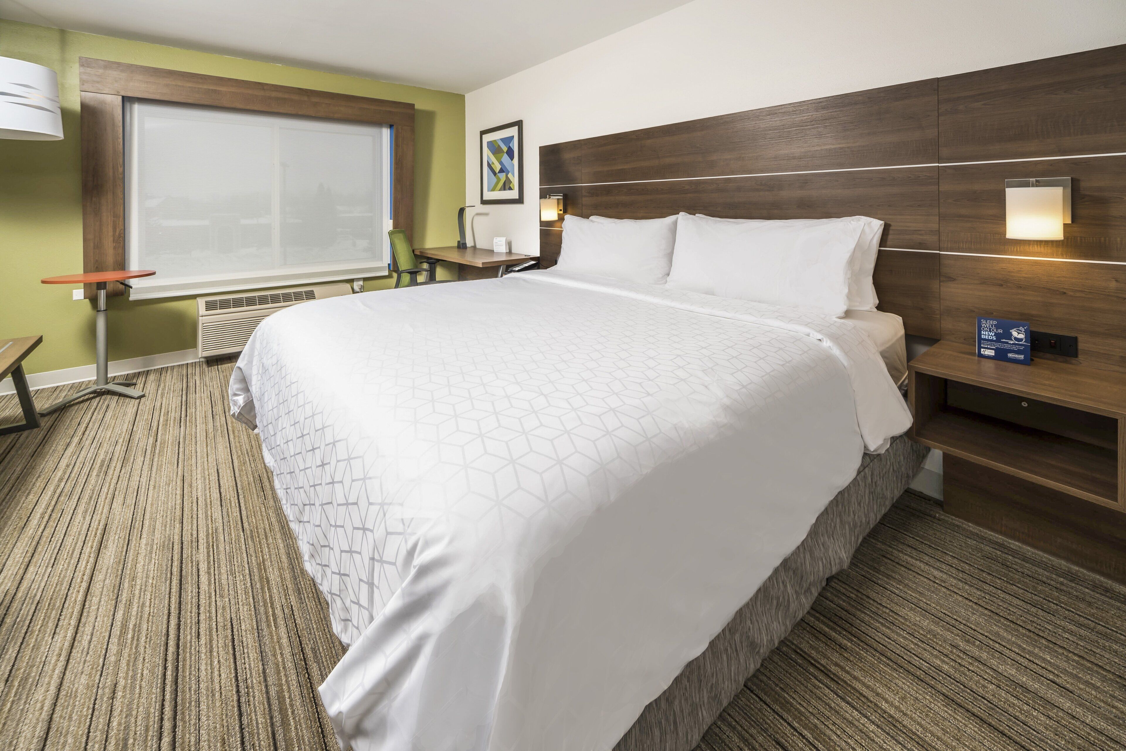 A hotel room with a large bed, a desk, a chair, a window with blinds, and a wall-mounted lamp.