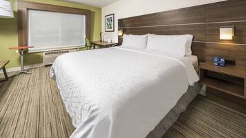 A hotel room with a large bed, a desk, a chair, a window with blinds, and a wall-mounted lamp.