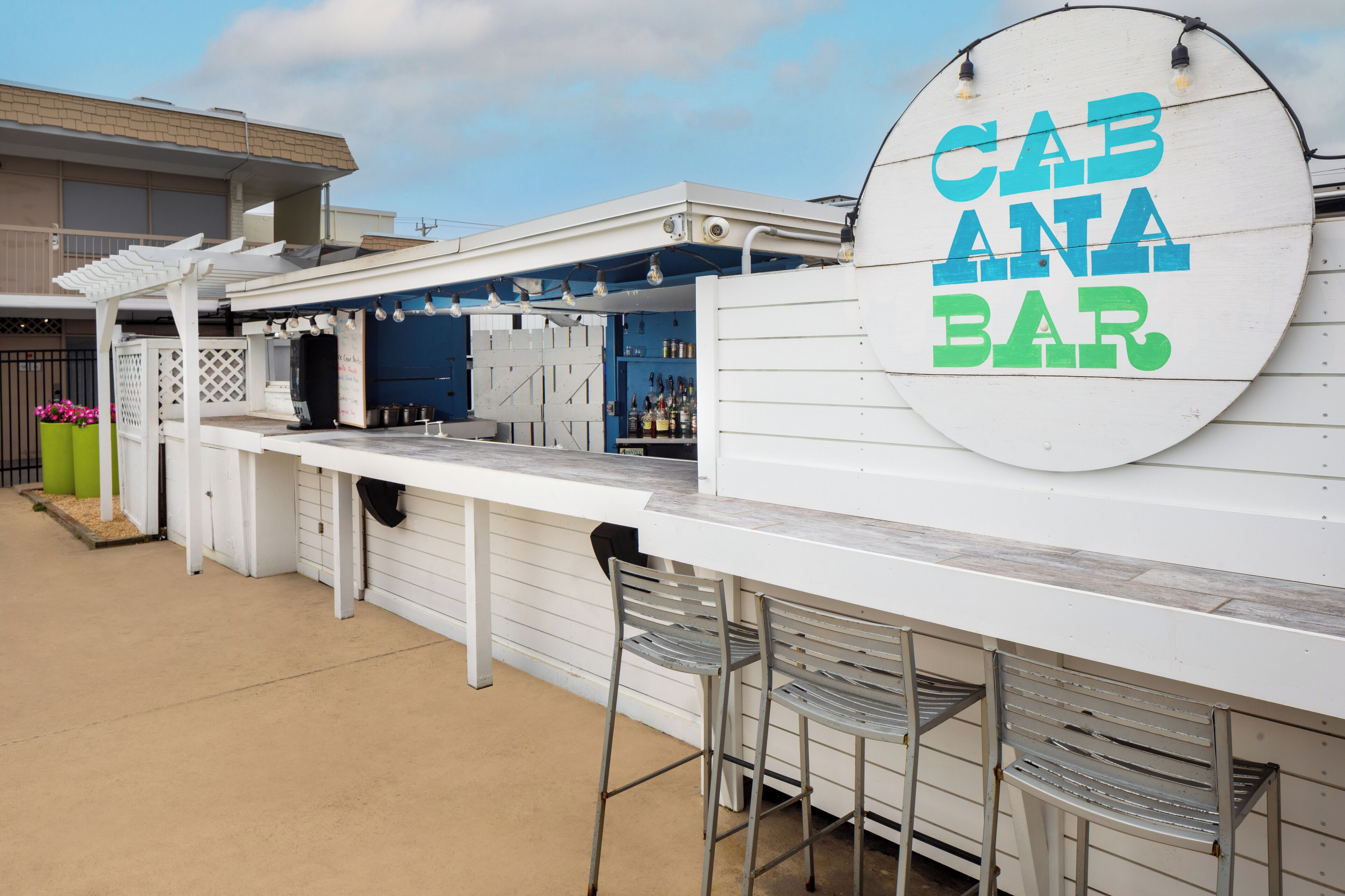 An outdoor bar area with a sign reading "CABANA BAR," featuring a white exterior, metal chairs, and a concrete floor.