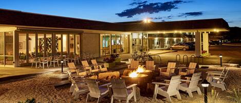 An outdoor seating area with lounge chairs and a fire pit, a building with a glass facade, and a parked car.