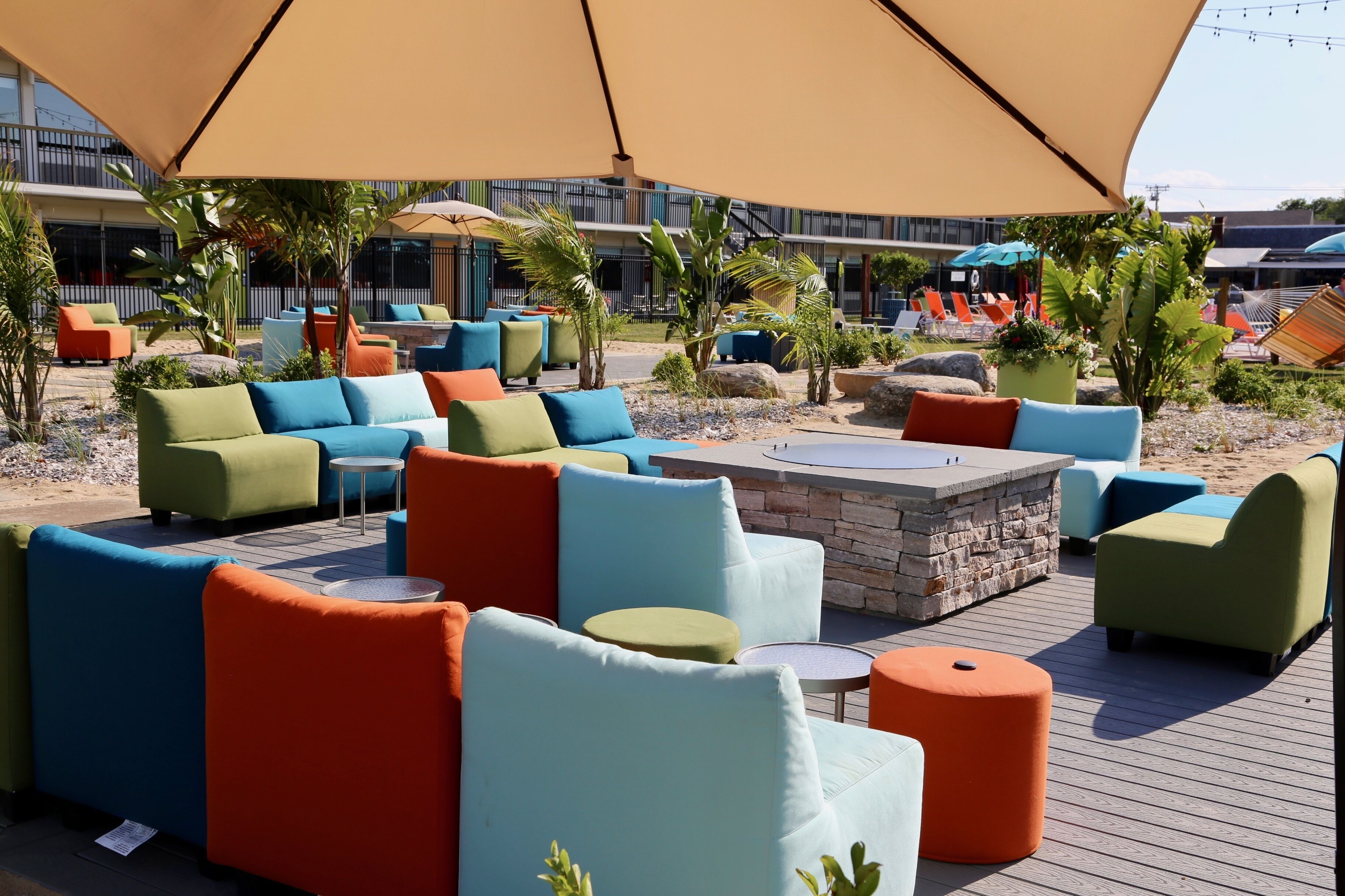 An outdoor seating area with colorful sofas and round tables, surrounded by palm trees and a large umbrella.