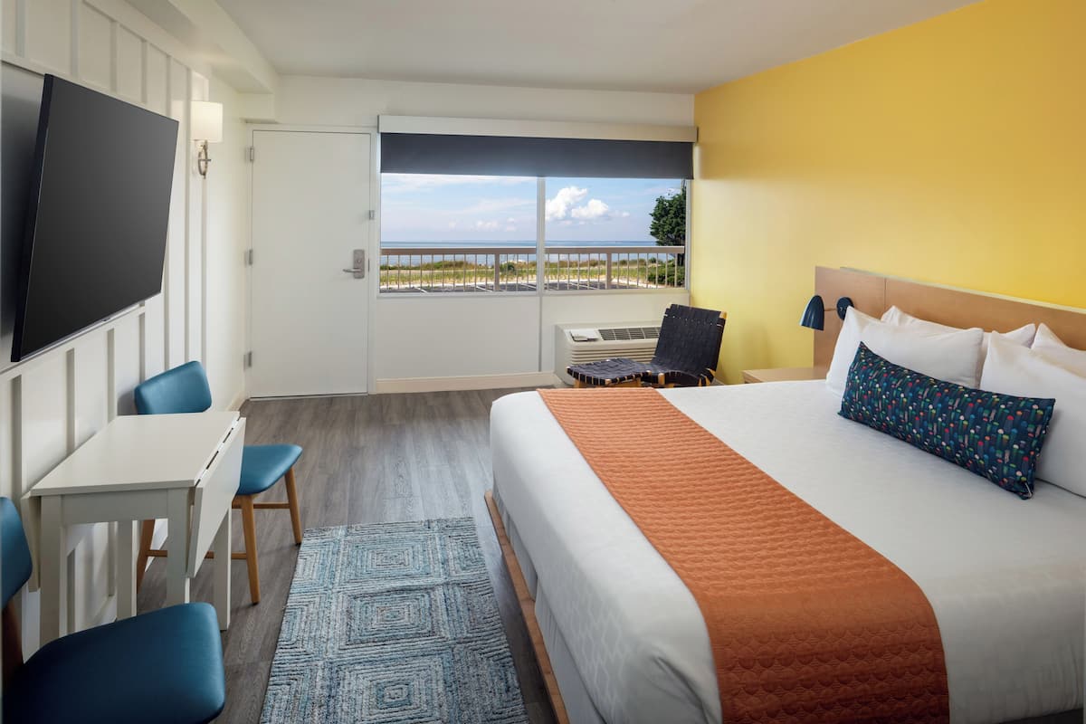 Une chambre d’hôtel équipée d’un lit, d’un bureau, d’une chaise, d’une télévision et d’une fenêtre donnant sur la plage.
