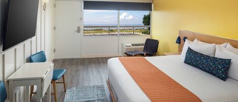 A hotel room with a bed, a desk, a chair, a TV, and a window with a view of the beach.