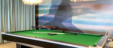 A pool table with a green felt surface, two chandeliers, and a mural of a mountain range on the wall behind it.