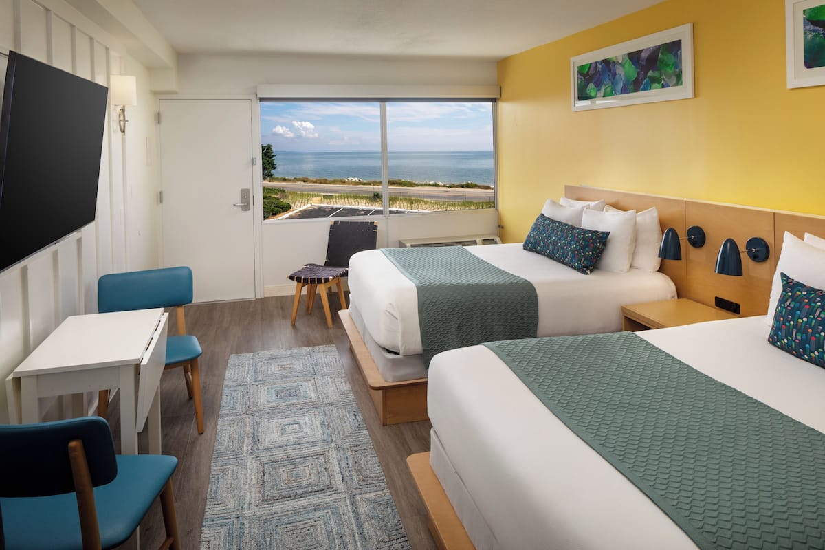 Une chambre d’hôtel avec deux lits, un bureau, des chaises, une télévision et une vue sur l’océan.
