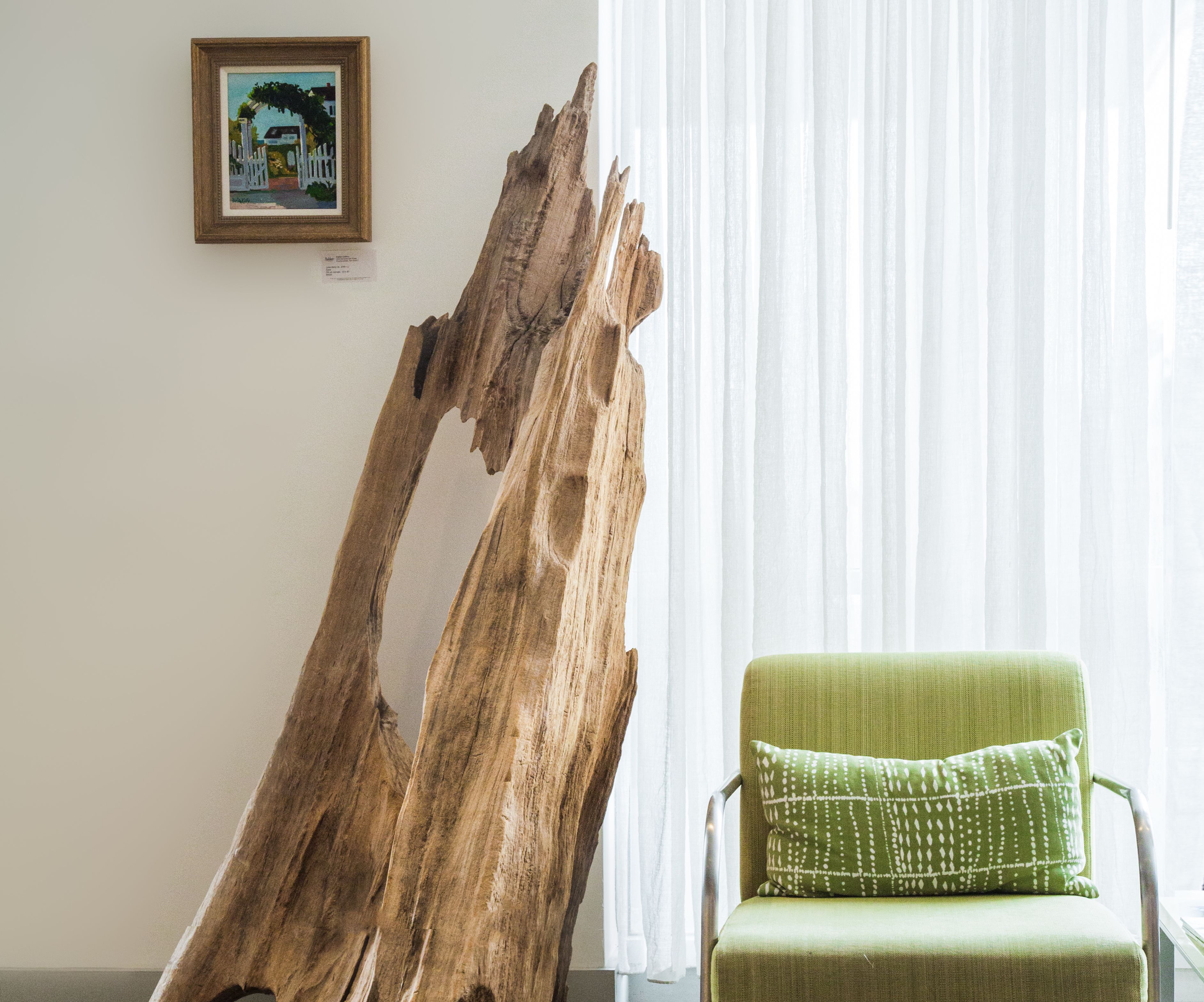 A room with a large wooden sculpture, a green armchair with a patterned pillow, and a framed picture on the wall.
