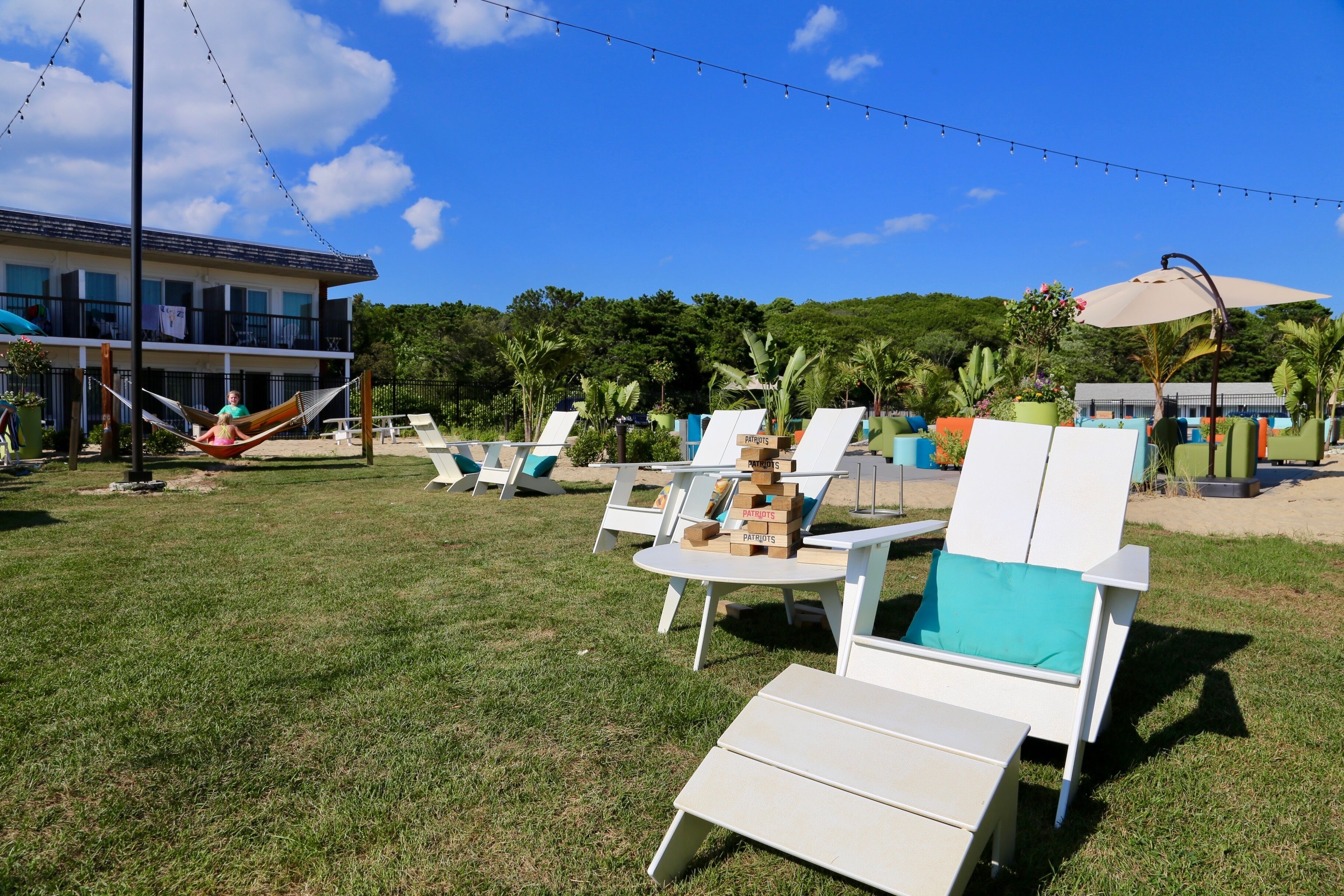 A grassy outdoor area with white lounge chairs and tables, a hammock, and string lights.