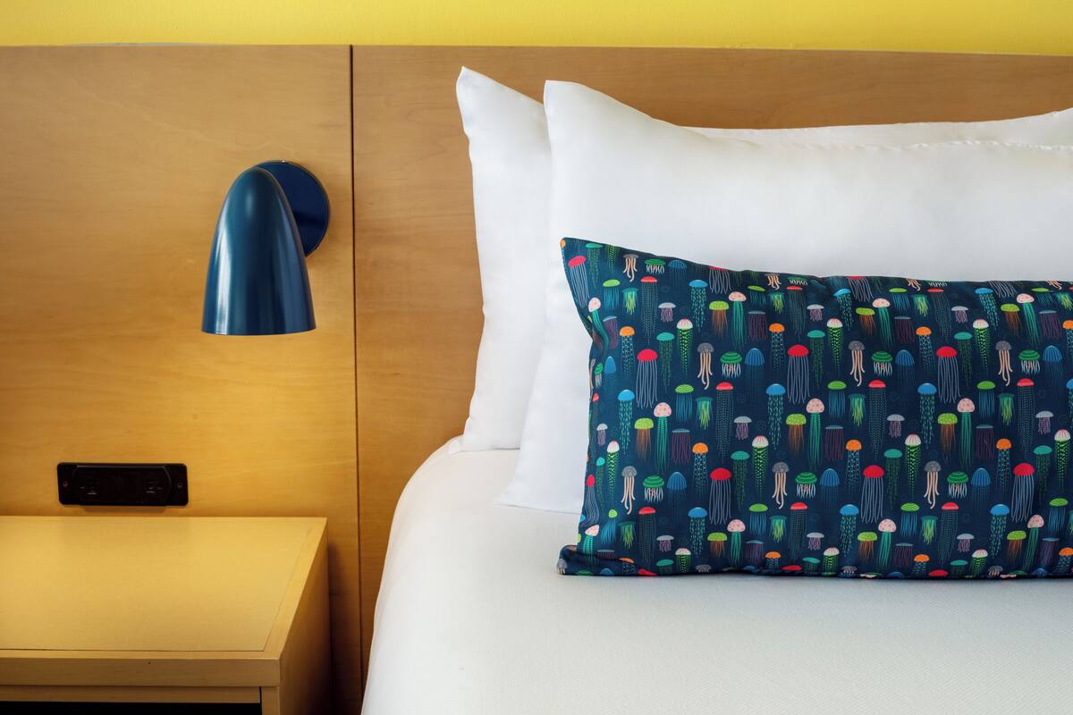Un lit bien fait, avec du linge de lit blanc et un coussin coloré, une table de chevet avec une lampe et une tête de lit en bois.