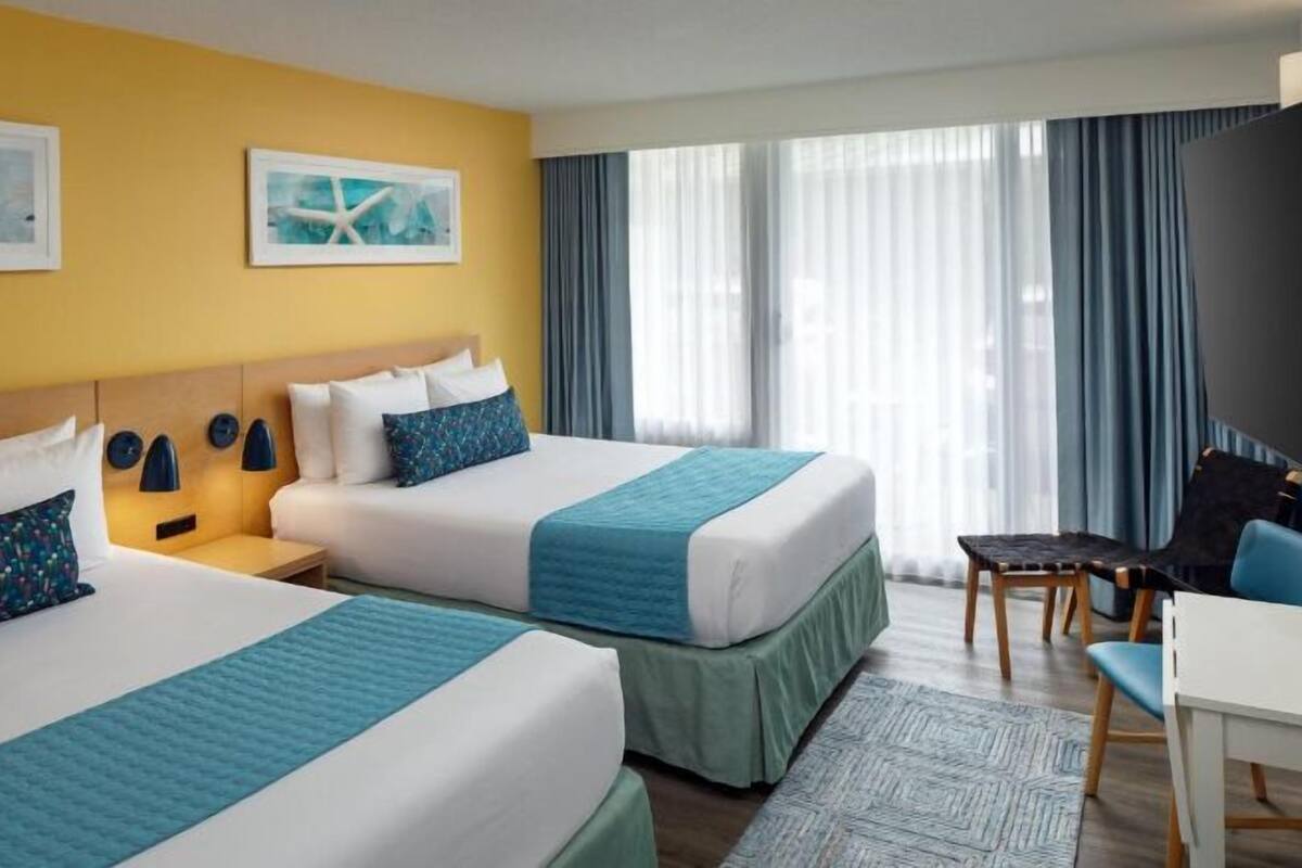 Une chambre d’hôtel avec deux lits, un bureau et une chaise. Il y a deux tableaux au mur, une fenêtre avec des rideaux et une vue sur l’extérieur.