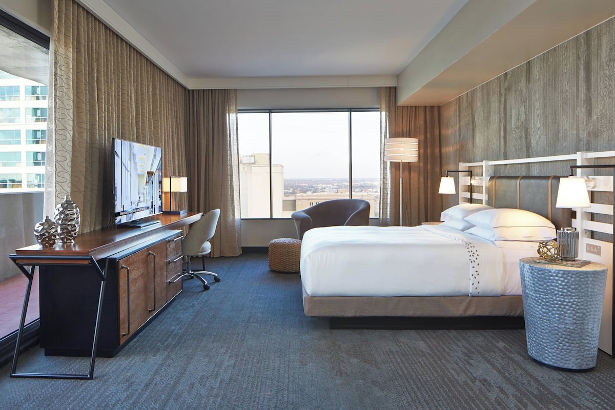 A hotel room with a bed, desk, chair, and a large window offering a city view.