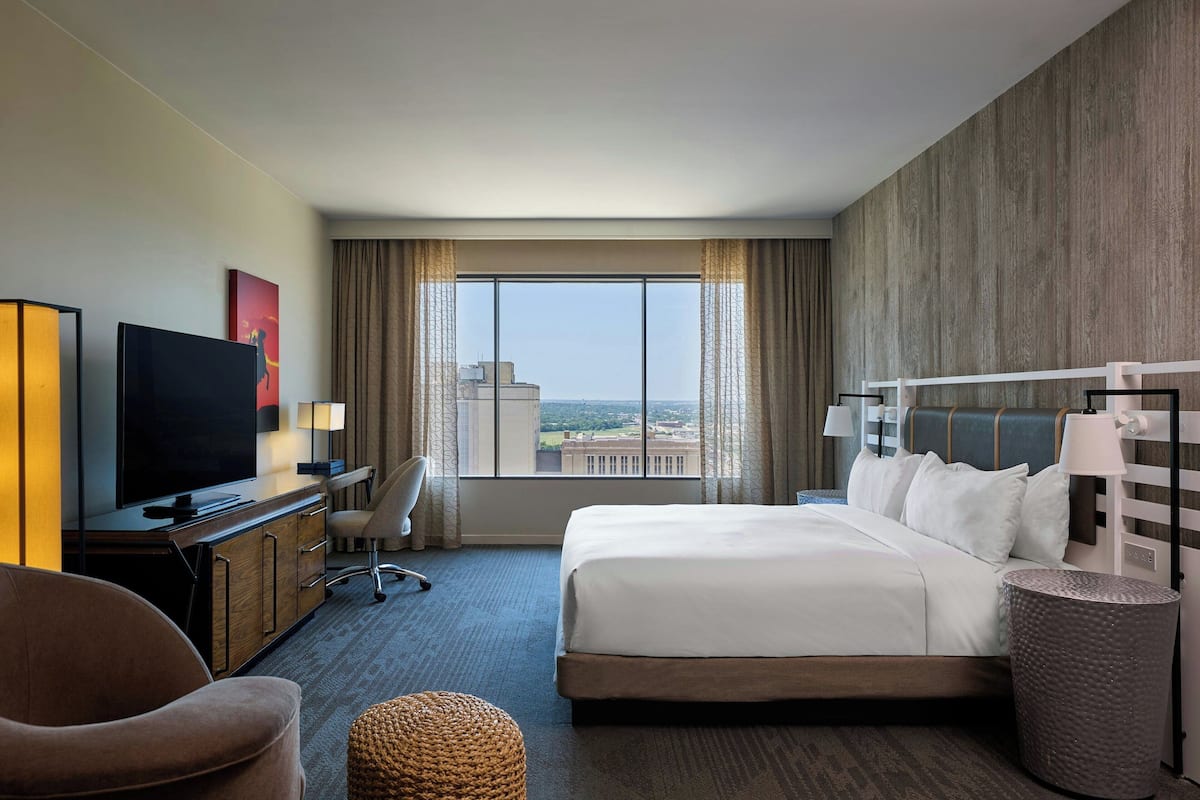 A hotel room with a large bed, a desk with a TV, a chair, a lamp, and a view of the city through the window.
