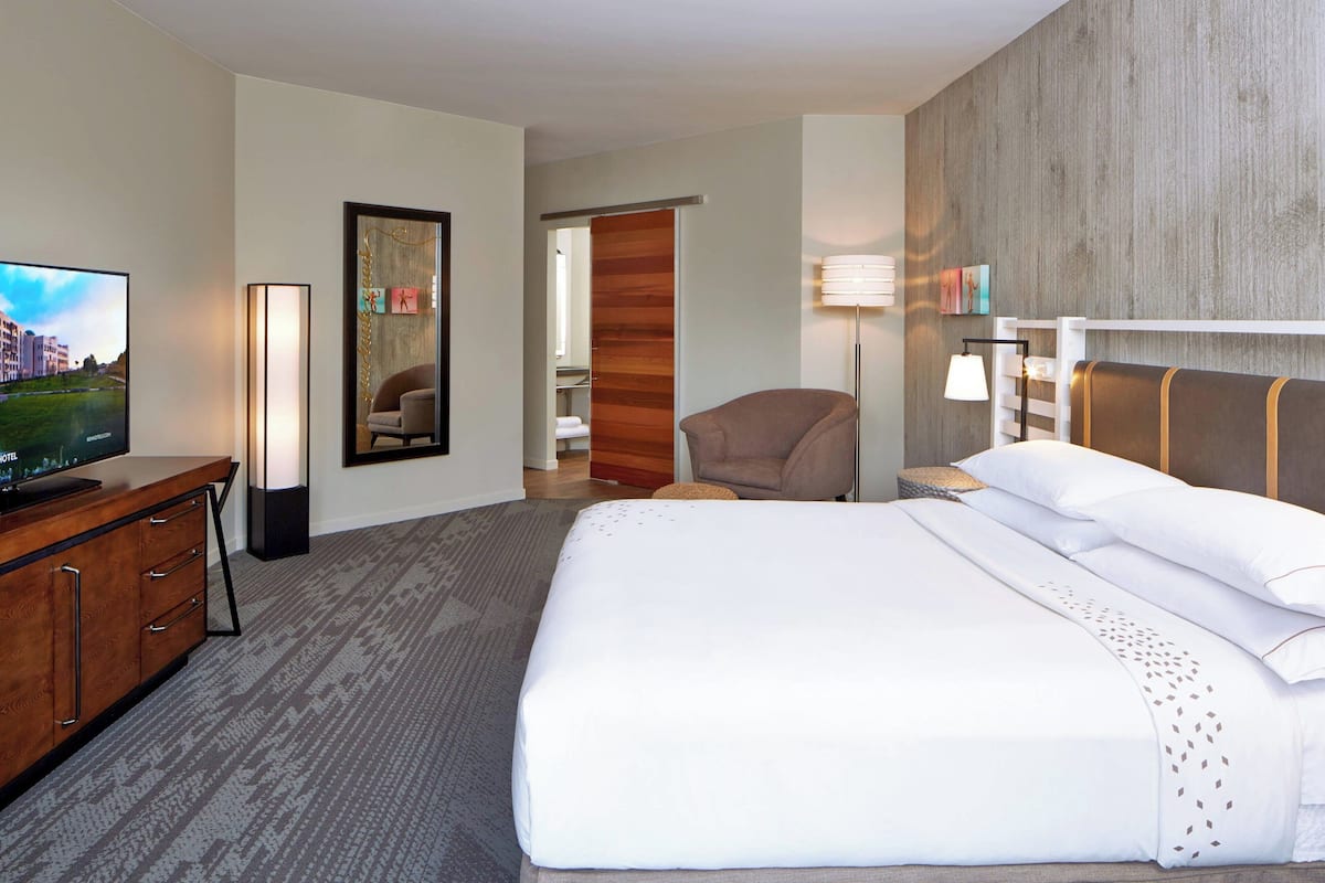 A hotel room with a large bed, a TV, a desk with drawers, a chair, a lamp, and a wooden wall panel.