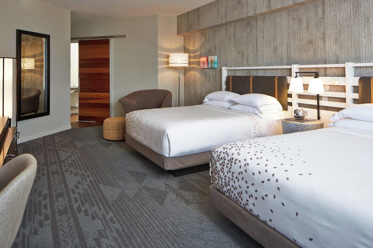 A hotel room with two beds, a chair, a lamp, and a mirror.