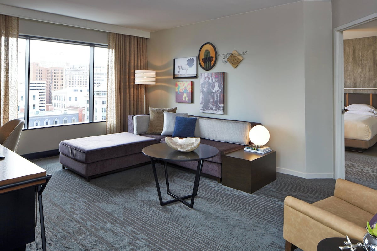 A hotel room with a large window, a sofa, a round table, a bed, and a nightstand with a lamp.