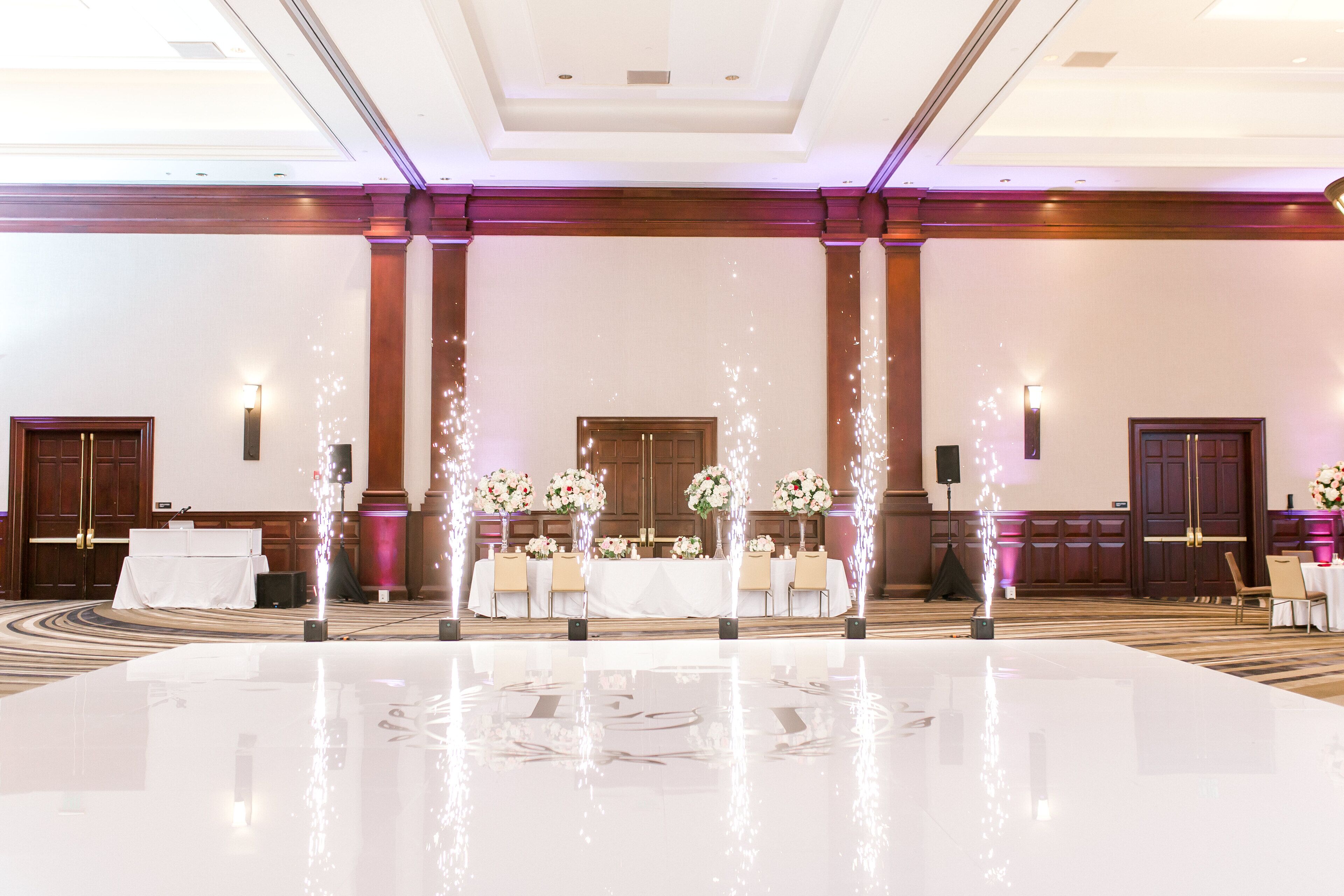 A spacious, well-lit banquet hall with a polished floor, a long table set for a formal event, and elegant floral arrangements.