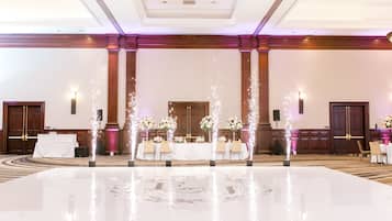 A spacious, well-lit banquet hall with a polished floor, a long table set for a formal event, and elegant floral arrangements.