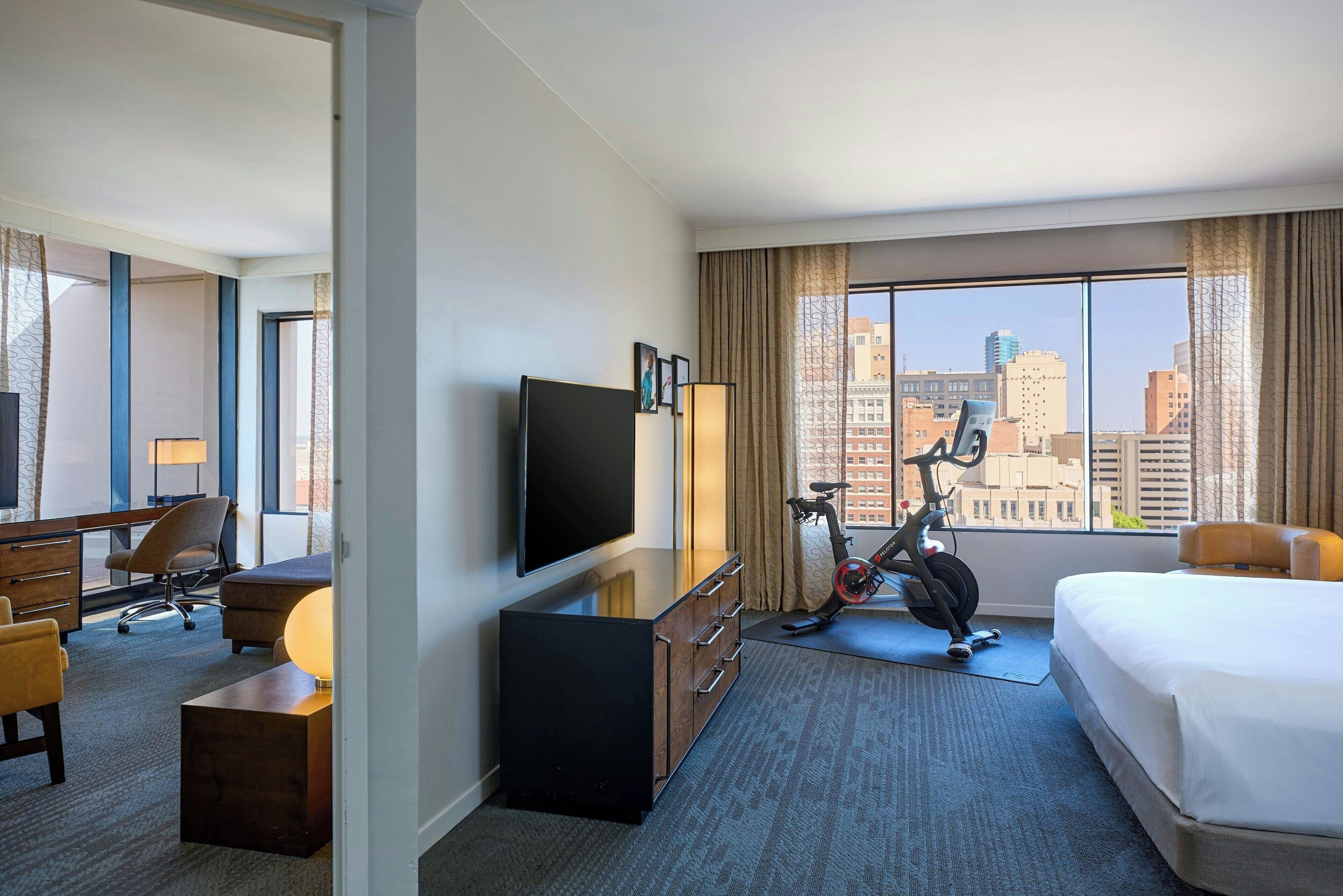 A hotel room with a bed, a desk with a chair, a TV, a lamp, an exercise bike, and a large window with a city view.