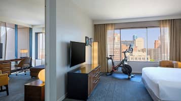 A hotel room with a bed, a desk with a chair, a TV, a lamp, an exercise bike, and a large window with a city view.