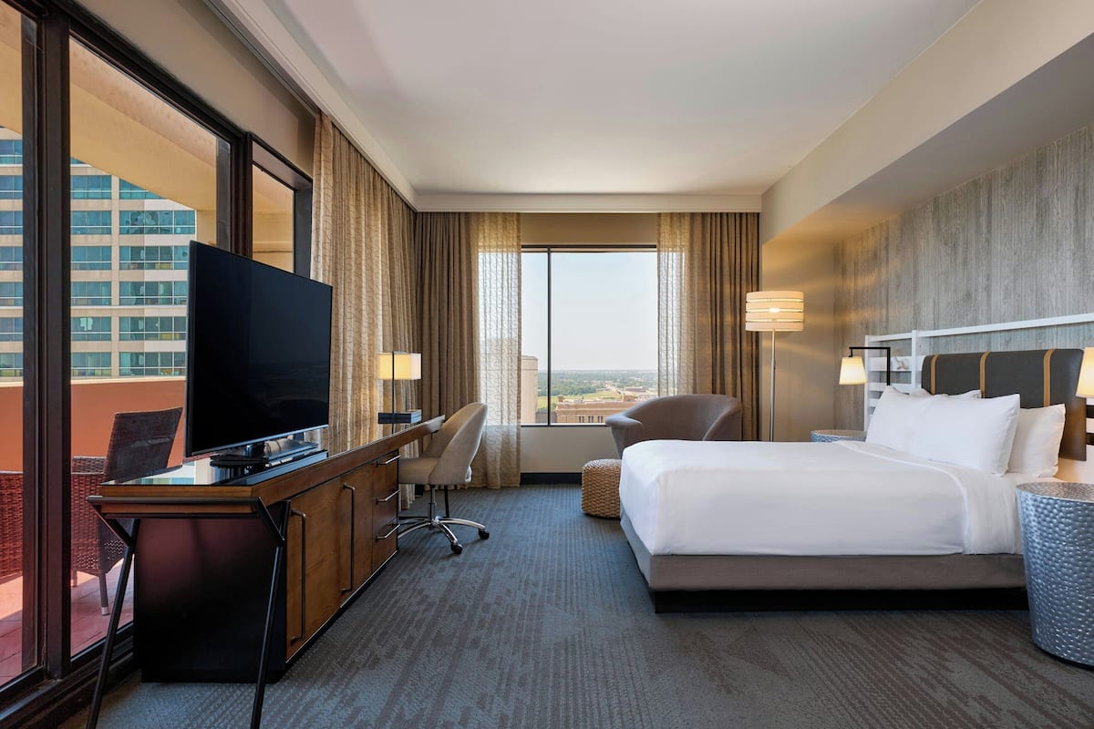 A hotel room with a large bed, a desk with a TV, a chair, and a view of a cityscape through large windows.