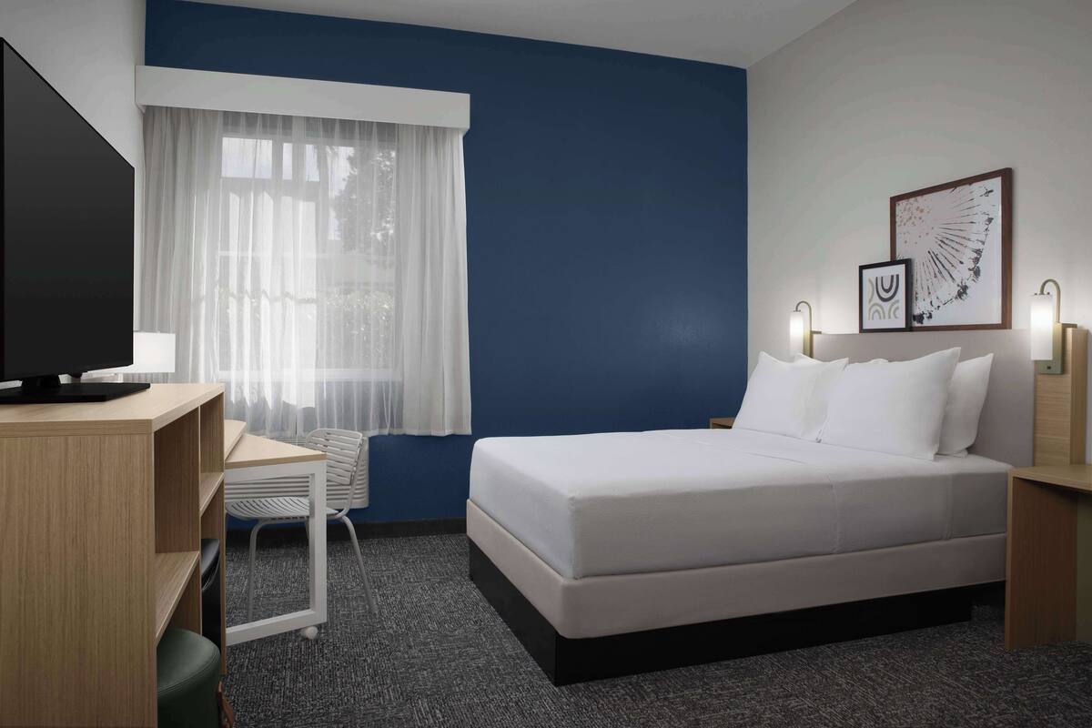 A hotel room with a bed, a desk, a chair, a television, and a window with sheer curtains.