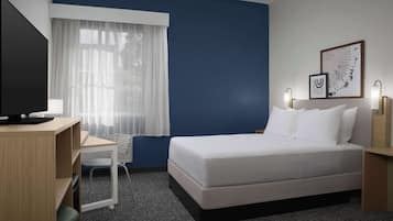 A hotel room with a bed, a desk, a chair, a television, and a window with sheer curtains.