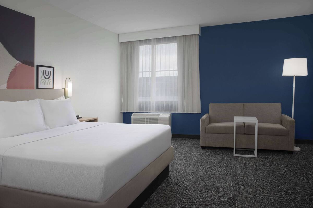 A hotel room with a large bed, a sofa, a small table, a window with curtains, and a wall-mounted air conditioner.