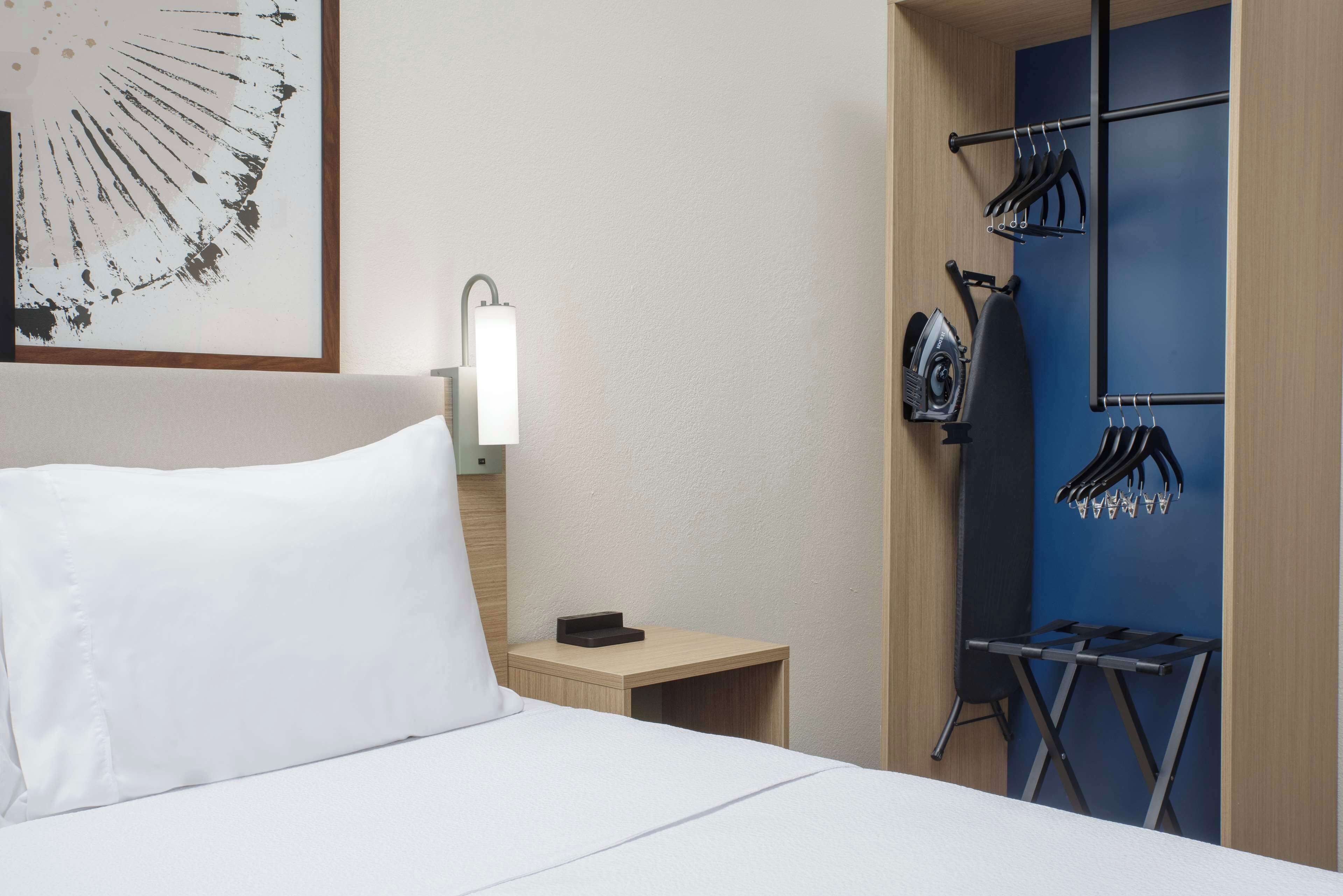 Habitación de hotel con cama, mesita de noche, tabla de planchar en la pared y un perchero con ropa.