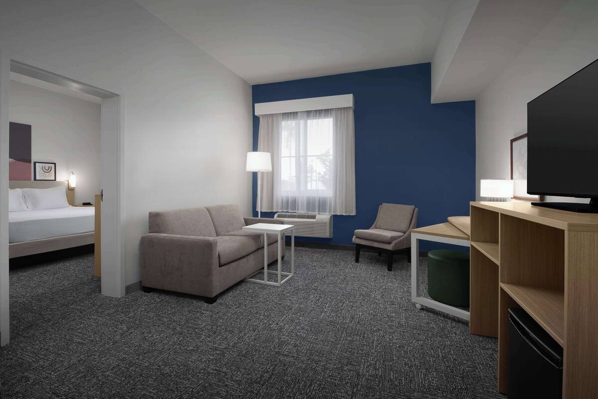 A hotel room with a bed, a sofa, a small table, a chair, a TV, and an air conditioning unit.