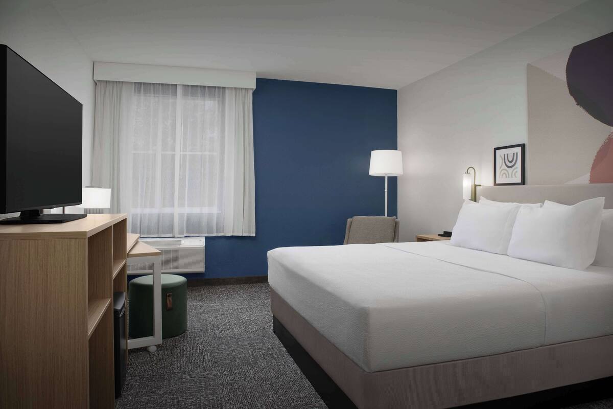 A hotel room with a large bed, a desk with a computer, a chair, a lamp, and a window with sheer curtains.
