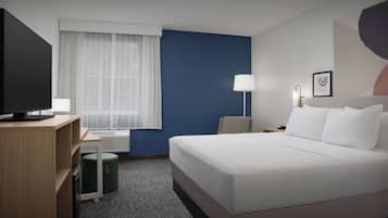 Habitación de hotel con una cama grande, un escritorio con computadora, una silla, una lámpara y una ventana con cortinas translúcidas.